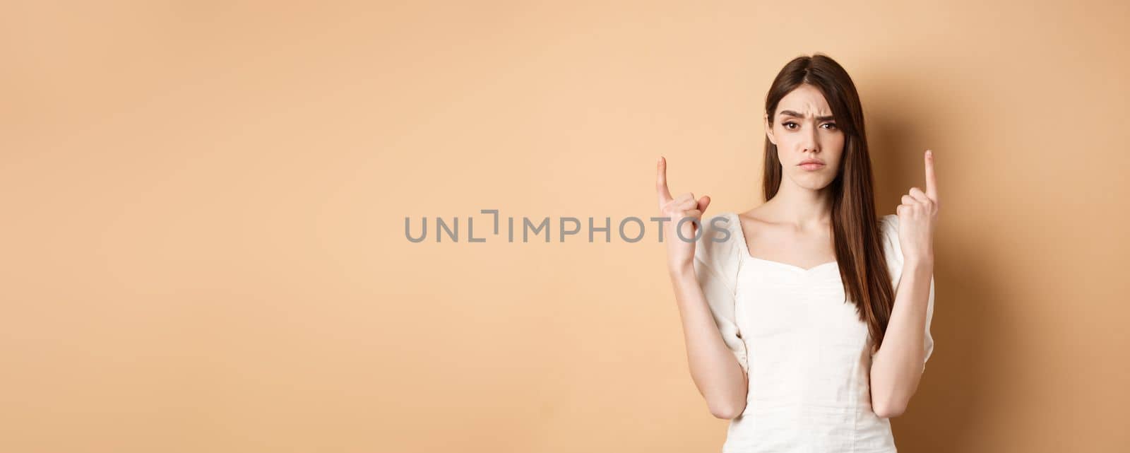 Skeptical woman frowning and pointing fingers up, having doubts about product, standing on beige background. Copy space