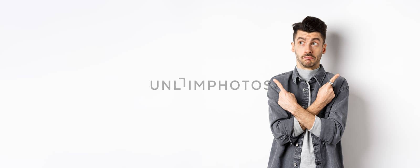 Indecisive handsome guy pointing fingers sideways, looking aside and making choice, showing two variants, standing unsure on white background.