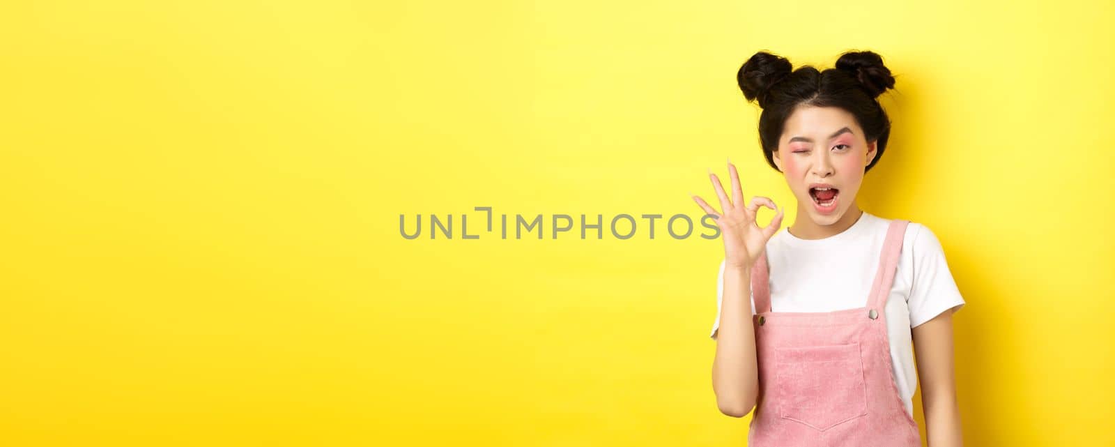 Alright. Confident asian female model with bright makeup and summer clothes, showing okay sign, winking and say yes, agree and approve, yellow background by Benzoix