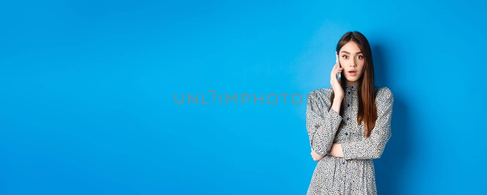 Shocked girl talking on mobile phone, gasping amazed at camera, speaking with someone and hear shocking news, standing in dress on blue background.