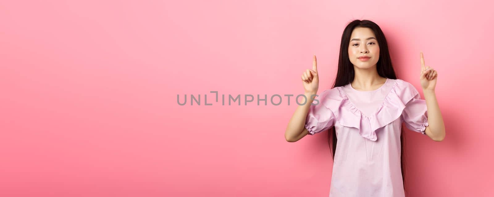 Smiling teen girl asian pointing fingers up at empty space, advertising on pink background by Benzoix