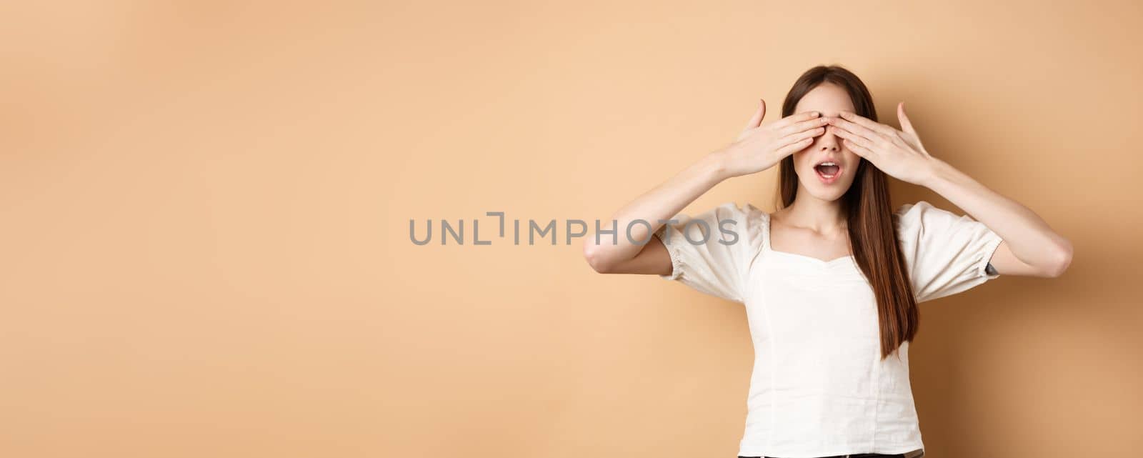 Happy romantic girl covering eyes and waiting for surprise, smiling while anticipating gift, standing on beige background.