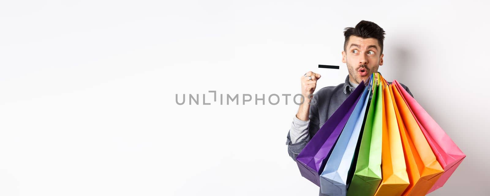 Excited young man buying in shop with plastic credit card, holding colorful shopping bags and looking aside at logo, standing against white background by Benzoix