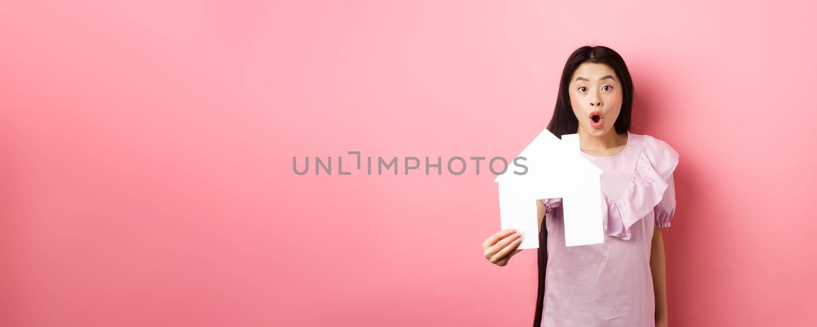Real estate and insurance concept. Excited asian woman showing paper house cutout and look amazed, special offer, standing on pink background.