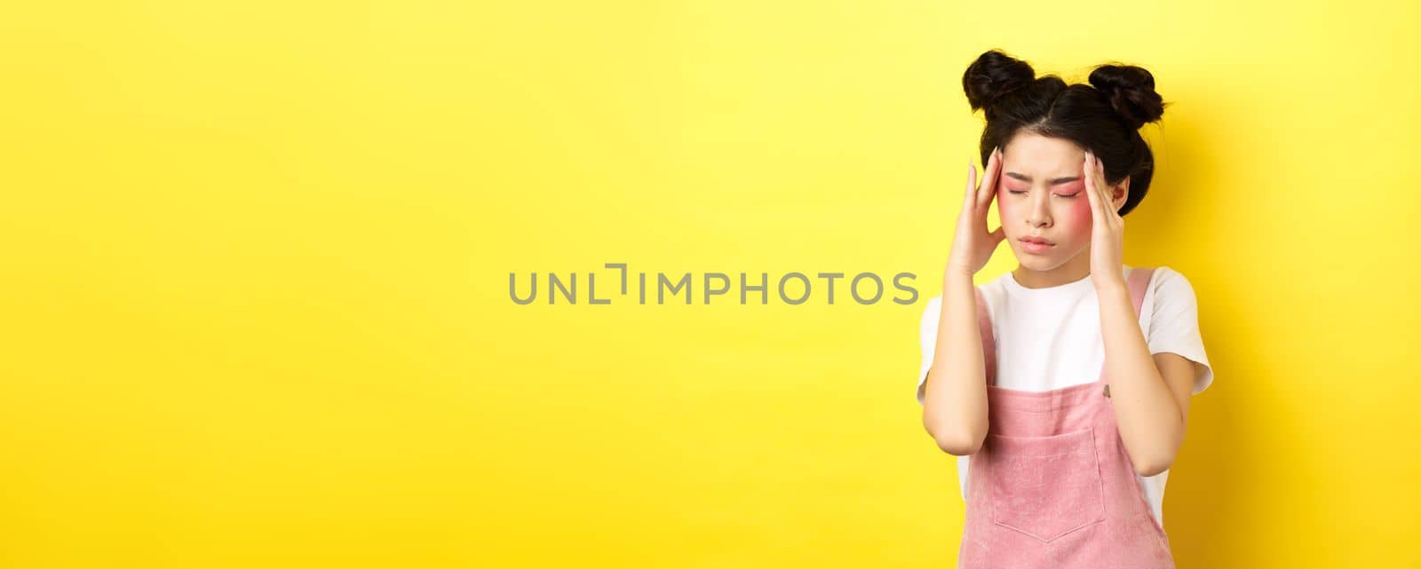 Image of stylish asian woman with bright makeup, having headache, touching head and looking tired, have painful migraine, yellow background.