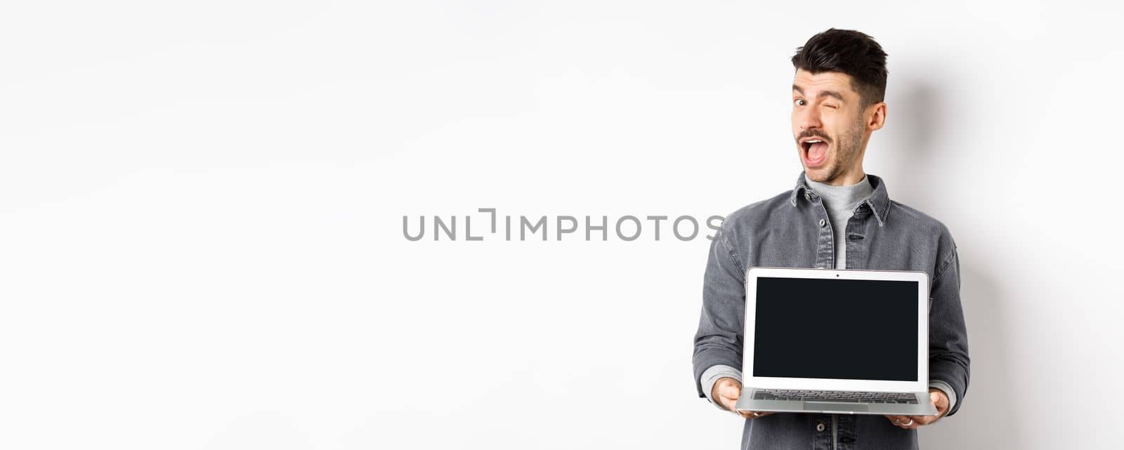 Cheerful guy winking and showing empty smartphone screen, offer you advertisement, standing on white background by Benzoix