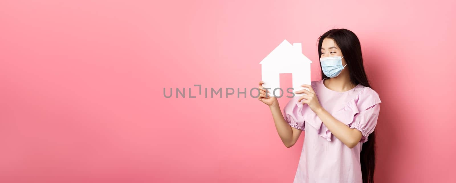 Real estate and pandemic concept. Excited asian girl in medical mask looking pleased at paper house cutout, buying property, standing on pink background.