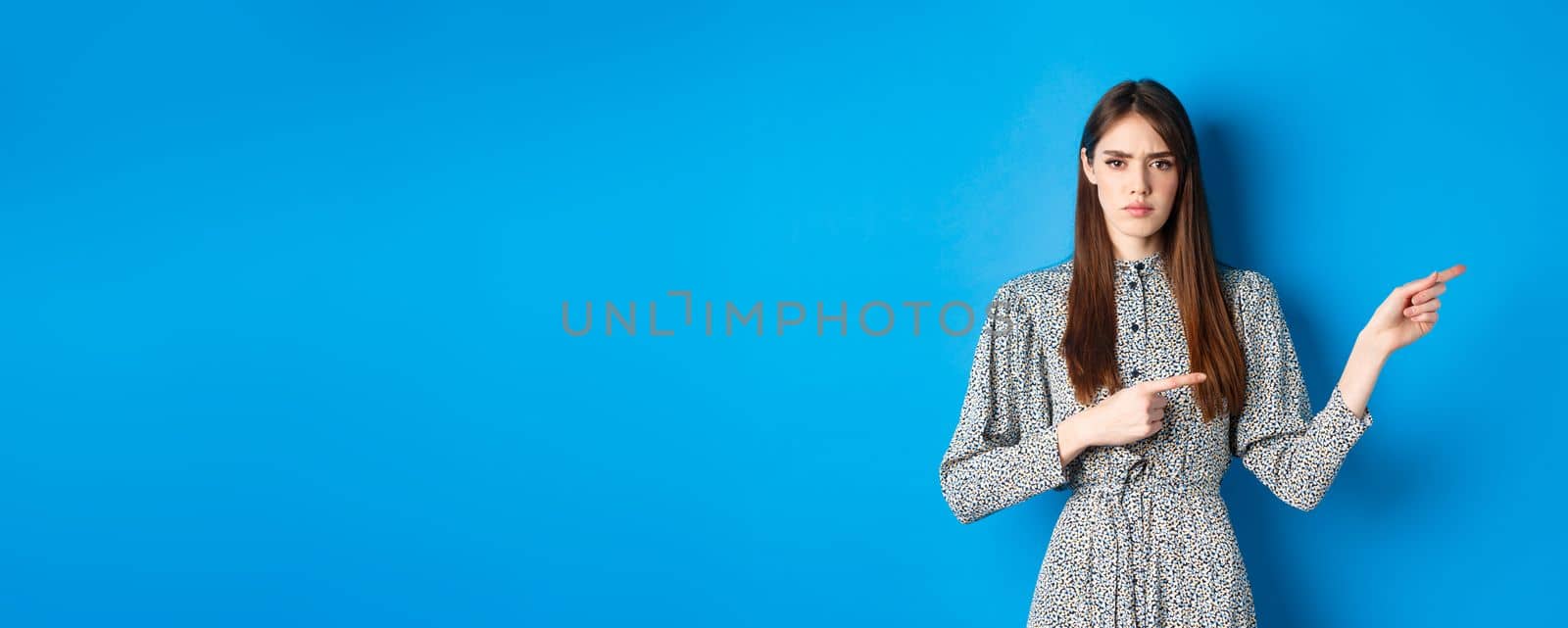 Angry frowning woman pointing fingers right and condemn person, feeling disapointed, standing in dress on blue background by Benzoix