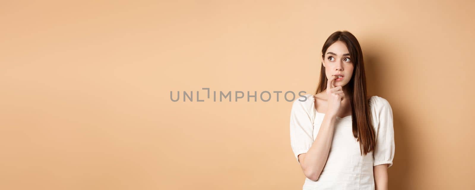 Nervous young girl biting fingernail and looking aside excited, making choice, thinking about something, standing on beige background.