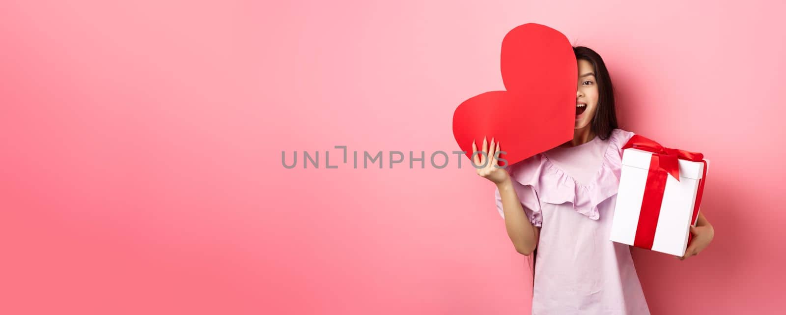 Valentines day concept. Happy young girl asian holding romantic gifts, red heart card from lover and present box, looking amazed at camera, standing in dress on pink background.