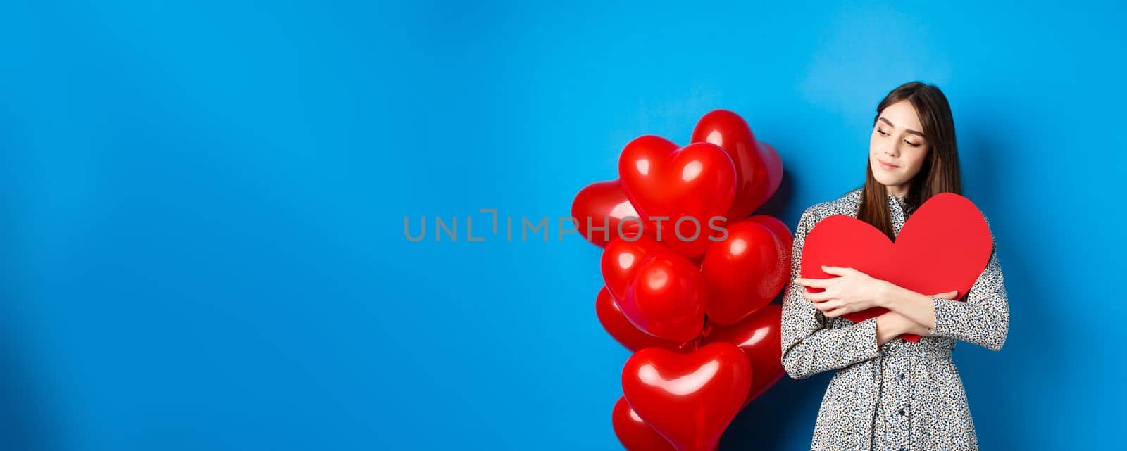 Valentines day. Romantic pretty woman in dress hugging big red heart cutout and looking dreamy, thinking of love, standing on blue background by Benzoix