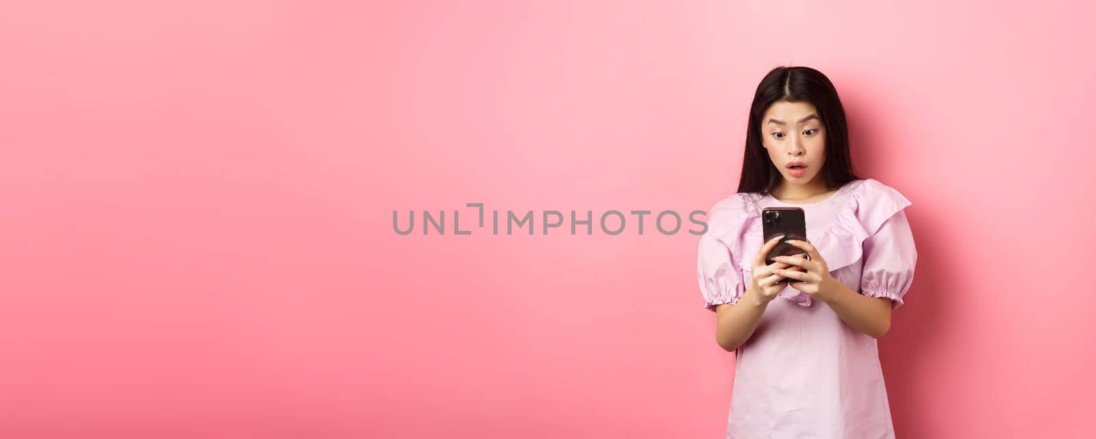 Online shopping. Asian teen girl look surprised at mobile phone screen, reading message with excited expression, standing against pink background.