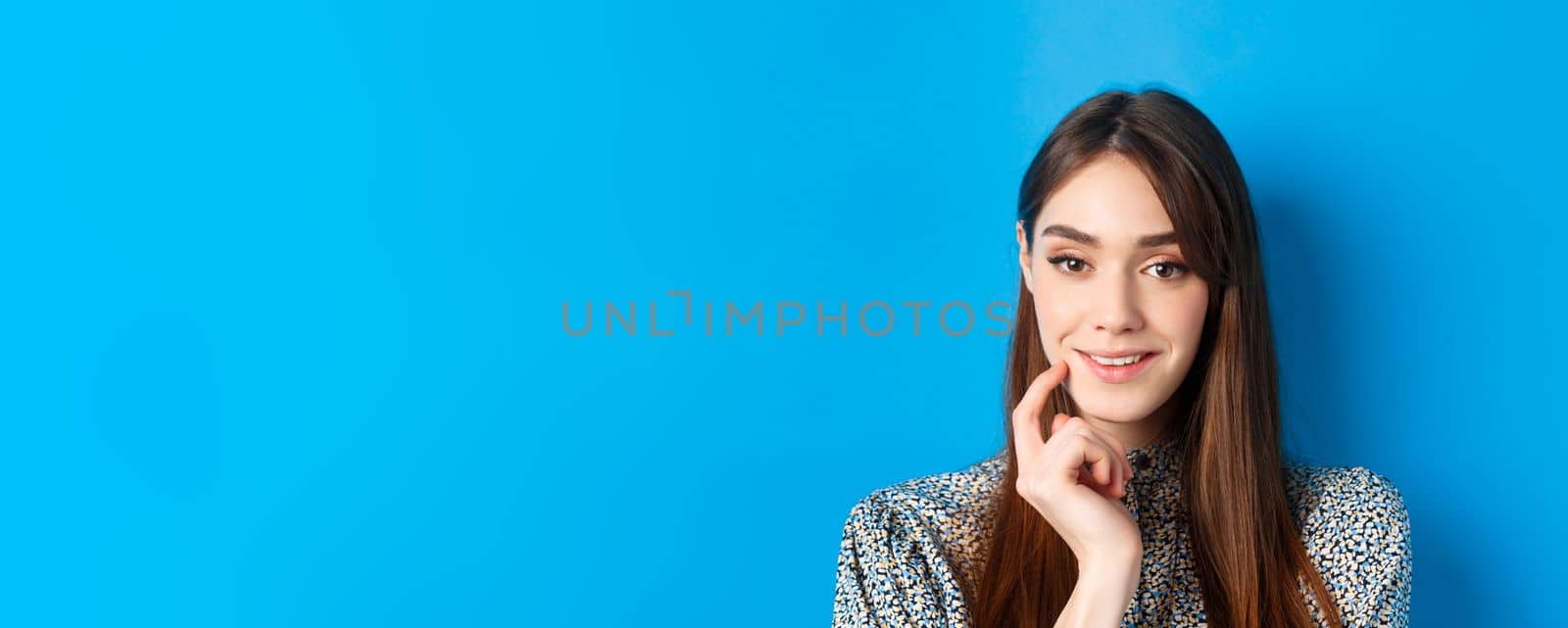 People emotions concept. Close-up portrait of intrigued pretty lady smiling and looking thoughtful, being interested, standing on blue background by Benzoix