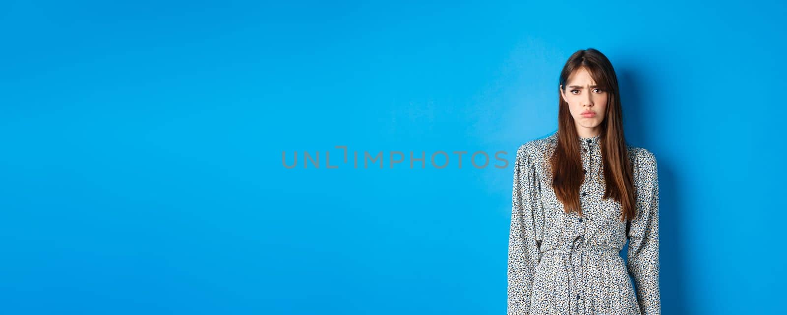 Moody frowning girl in dress looking at unfair thing, standing offended or upset against blue background by Benzoix