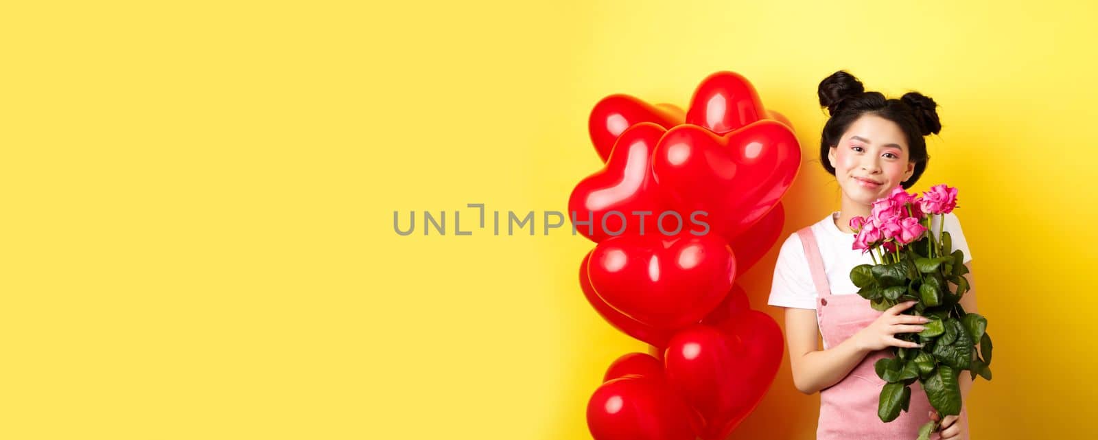 Happy Valentines day. Beautiful asian woman dressed for romantic date, holding bouquet of flowers and smiling. Girl with roses standing near heart balloons, yellow background.
