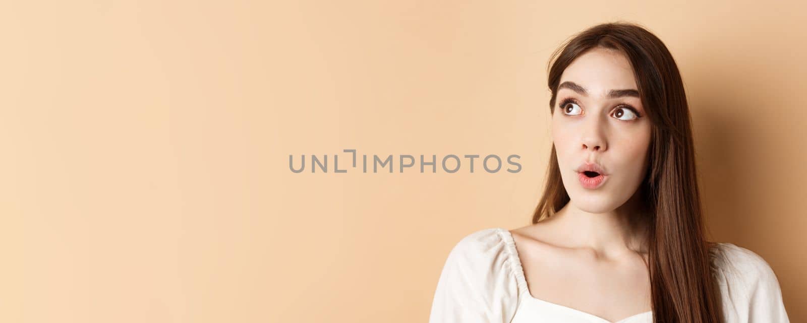 Close up portrait of excited pretty woman looking aside logo, say wow with amazed face, standing on beige background by Benzoix