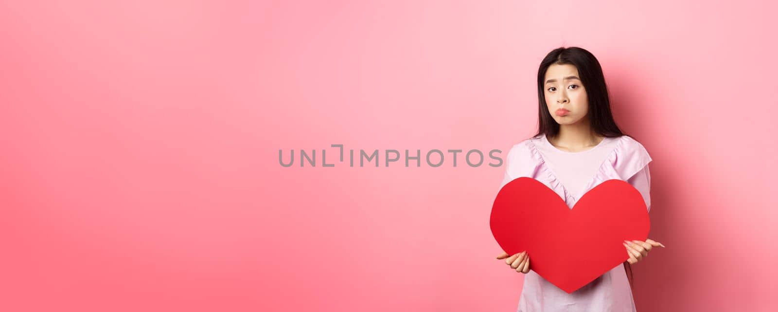 Valentines concept. Single teenage asian girl wants to fall in love, looking sad and lonely at camera, sulking distressed on lovers day, holding big red heart cutout, pink background.