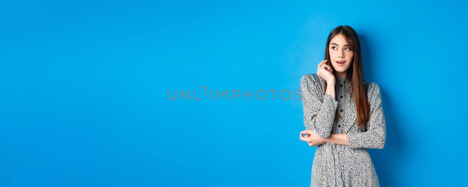 Pensive caucasian girl with long natural hair, wearing vintage dress, looking left with thoughtful face, standing on blue background by Benzoix