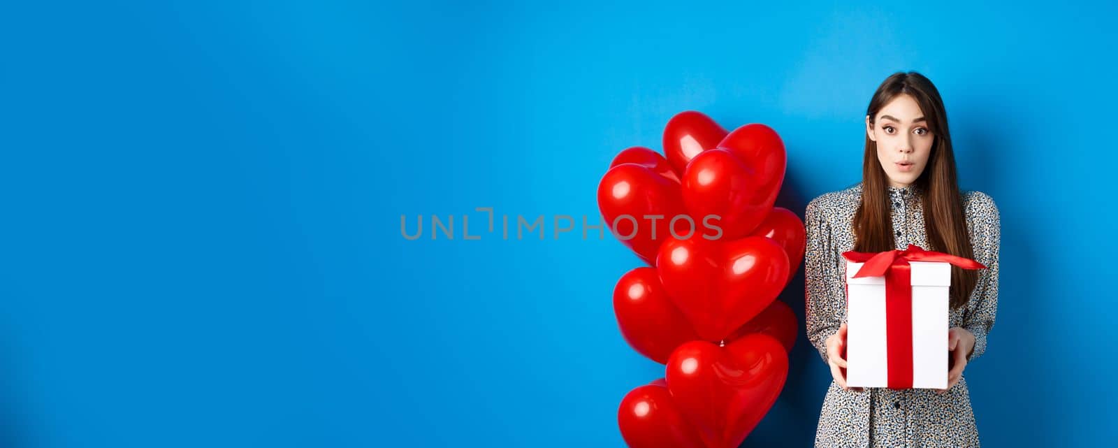 Valentines day. Surprised attractive girl looking amazed at camera, holding big romantic gift, standing near red hearts balloons, blue background.