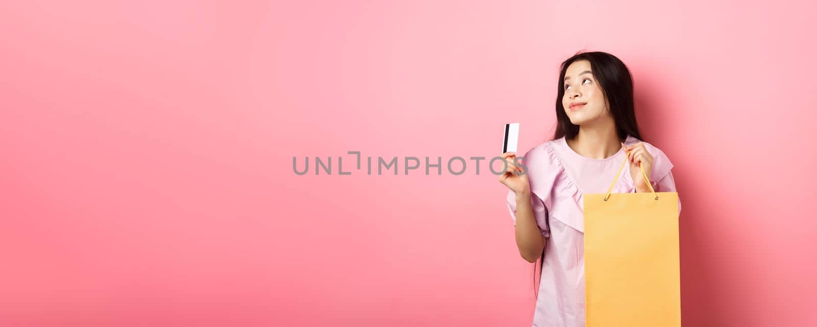 Shopping. Dreamy asian girl thinking of buying something, holding shop bag and plastic credit card, standing against pink background by Benzoix