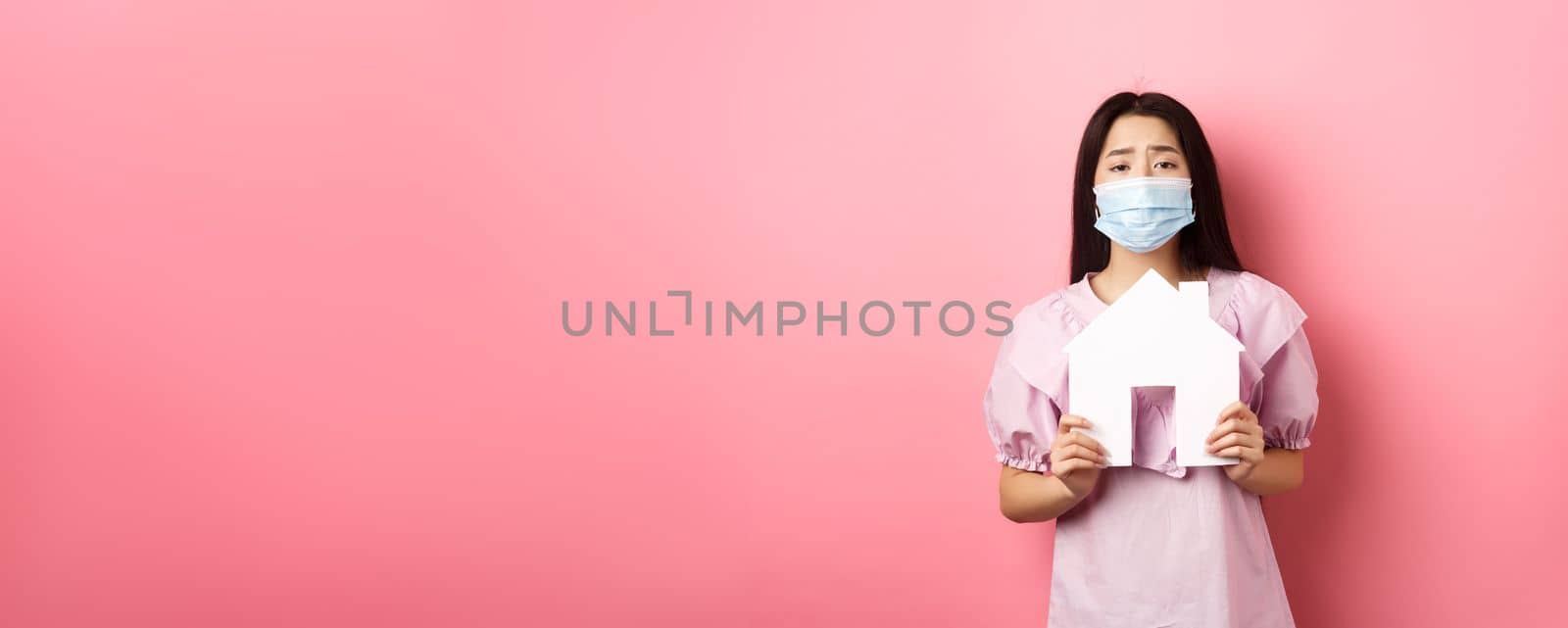 Real estate and pandemic concept. Begging asian girl showing paper house cutout, wearing medical mask, asking for apartment, standing against pink background by Benzoix