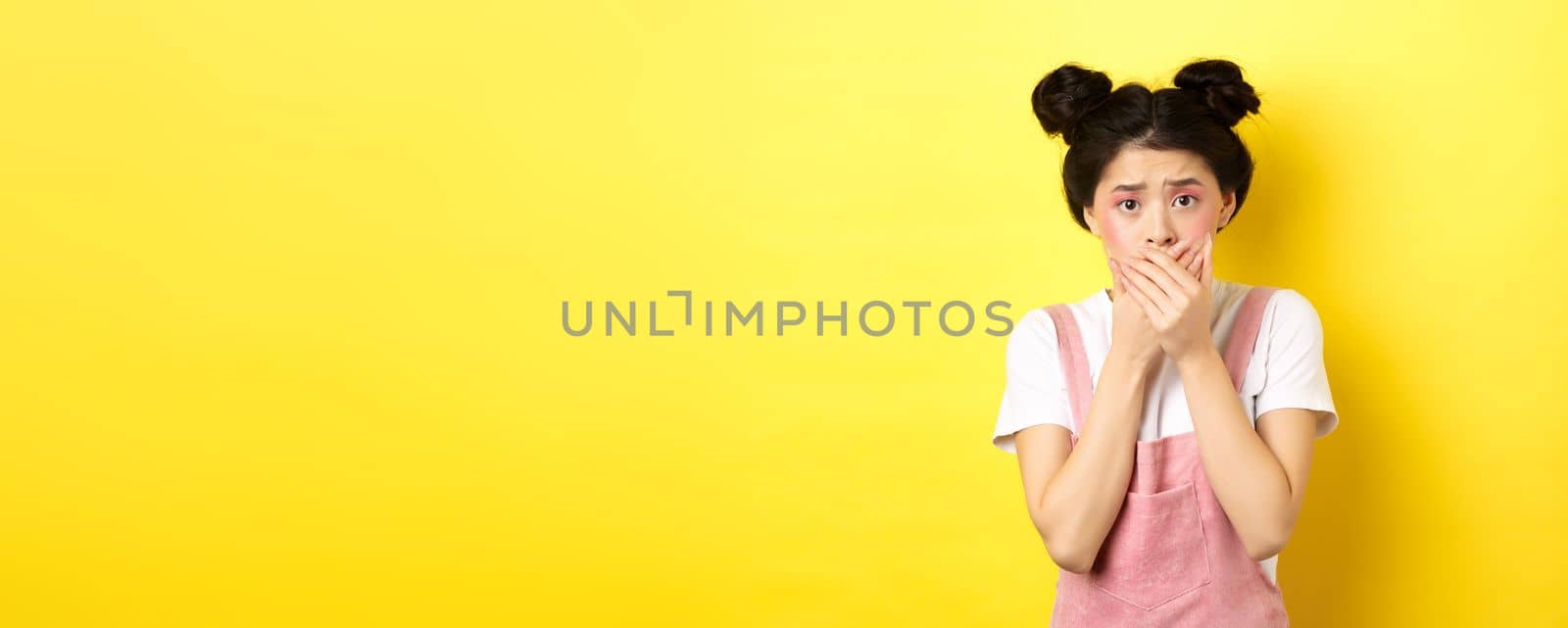 Worried asian teen girl covering mouth with hands, looking concerned and anxious, standing scared on yellow background with glam makeup and summer clothes.