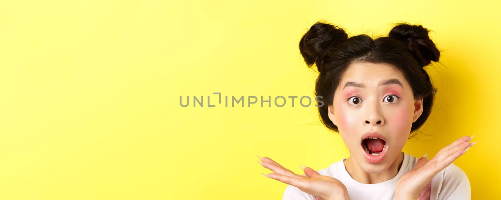 Close-up of excited asian woman with glamour makeup and hairstyle, screaming of amazement, open mouth and look at camera in awe, yellow background.