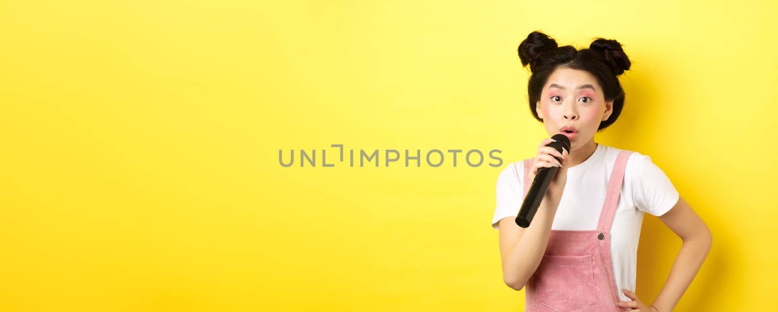 Cute asian teen girl with bright makeup, singing in microphone karaoke, standing against yellow background.