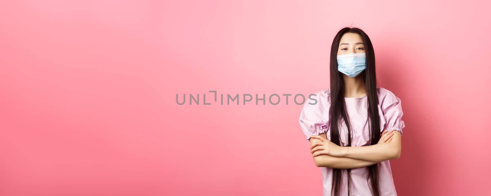 Covid-19, pandemic lifestyle concept. Cute asian woman in medical mask looking confident, cross arms on chest in self-assured pose, standing against pink background by Benzoix