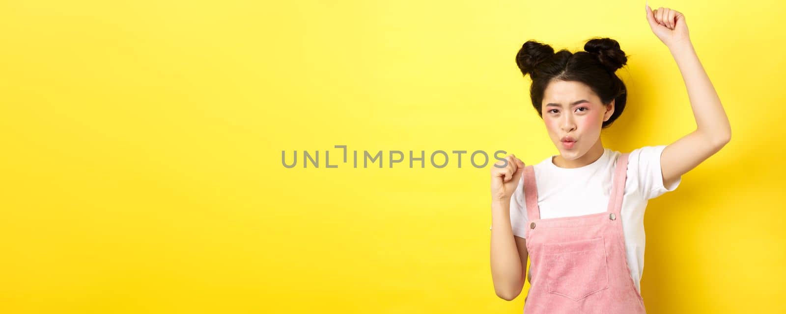 Excited asian woman getting motivation, raising hand up and chanting, celebrating victory, triumphing and standing on yellow background.