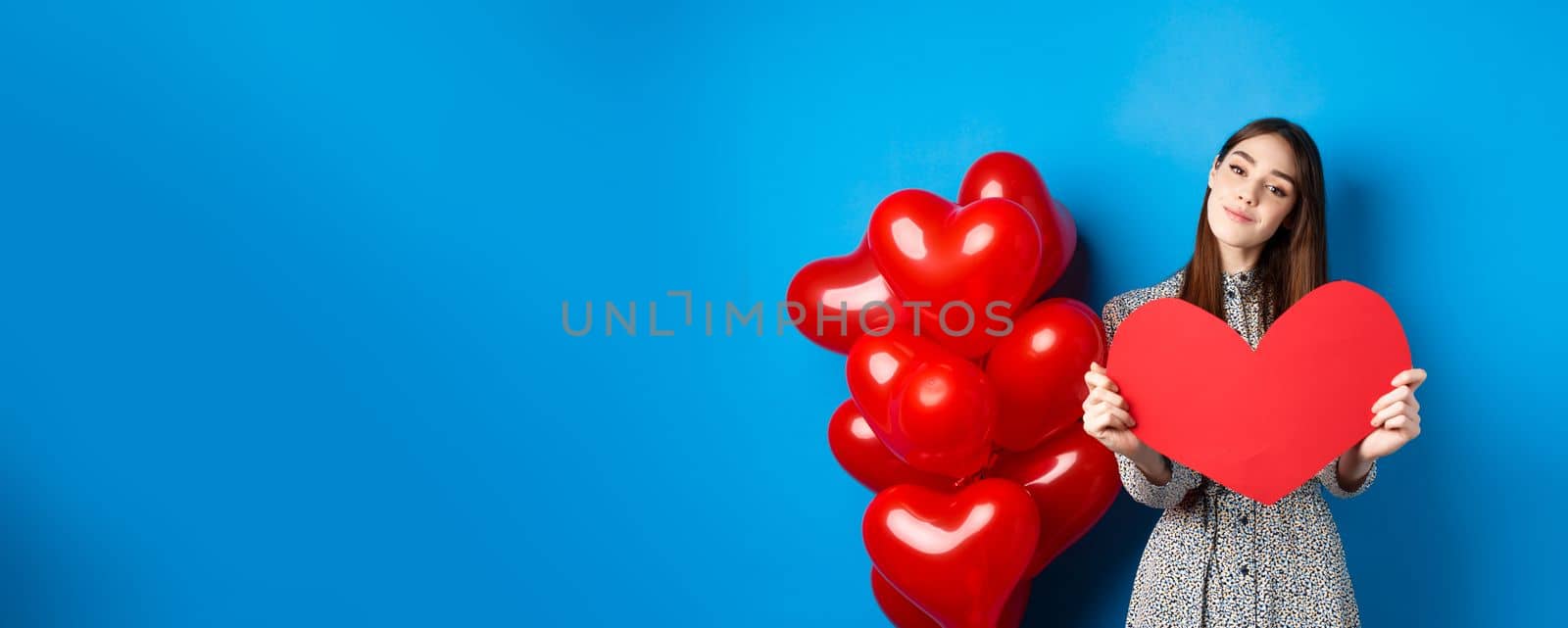Valentines day. Romantic girl in dress showing big red heart cutout, dreaming of love, standing near holiday balloons on blue background by Benzoix