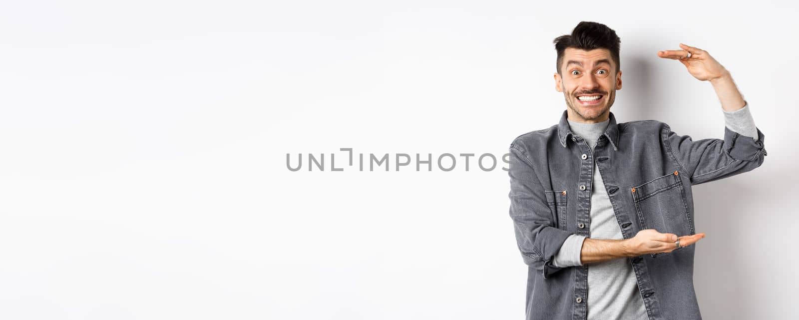 Cheerful guy smiling and showing big size, showing large thing and looking excited, standing against white background.