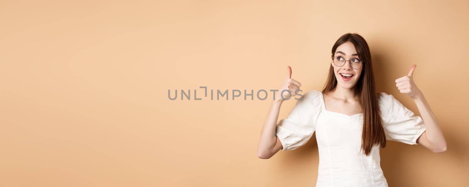 Happy girl in glasses show thumbs up, looking aside at good logo, standing satisfied on beige background by Benzoix