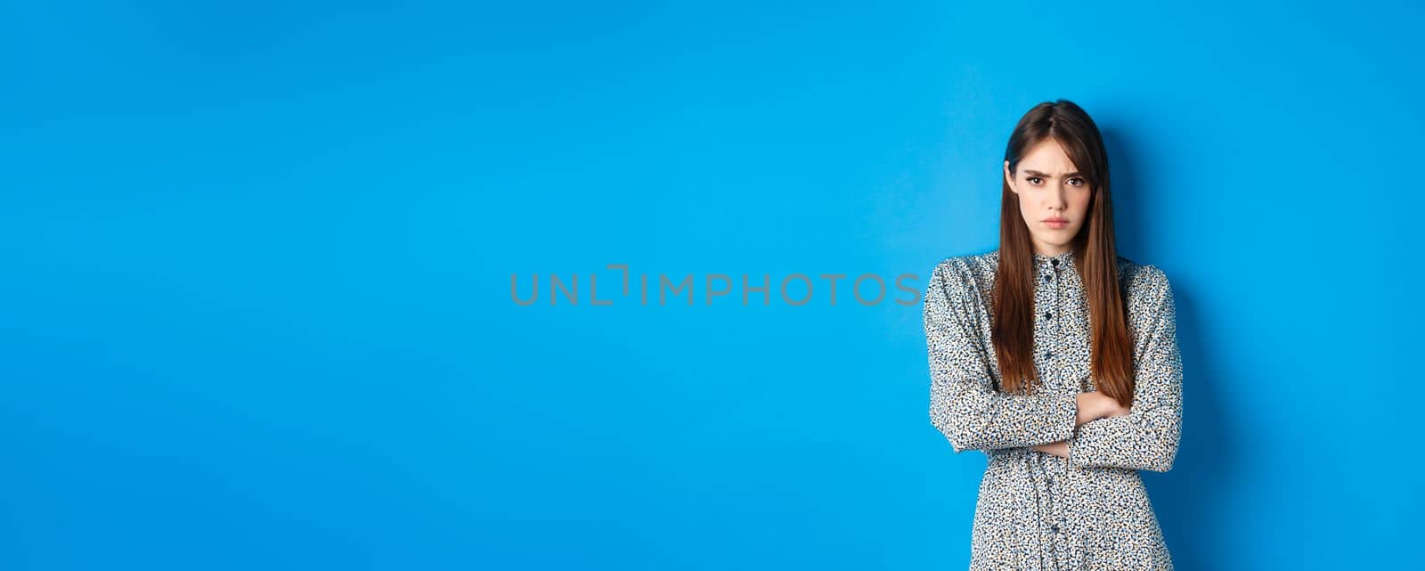 Angry frowning woman in dress, cross arms on chest and feel offended about something unfair, standing upset against blue background by Benzoix