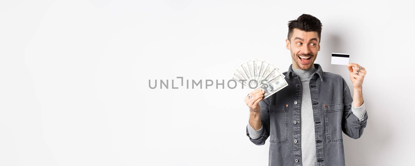 Excited guy holding plastic credit card and dollar bills, standing amused on white background by Benzoix