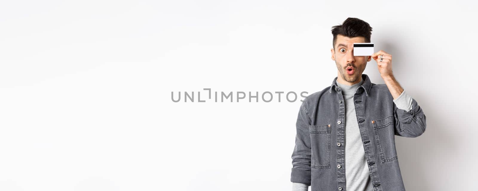 Excited funny man hold plastic credit card on eye and say wow, checking out special deal, standing on white background.