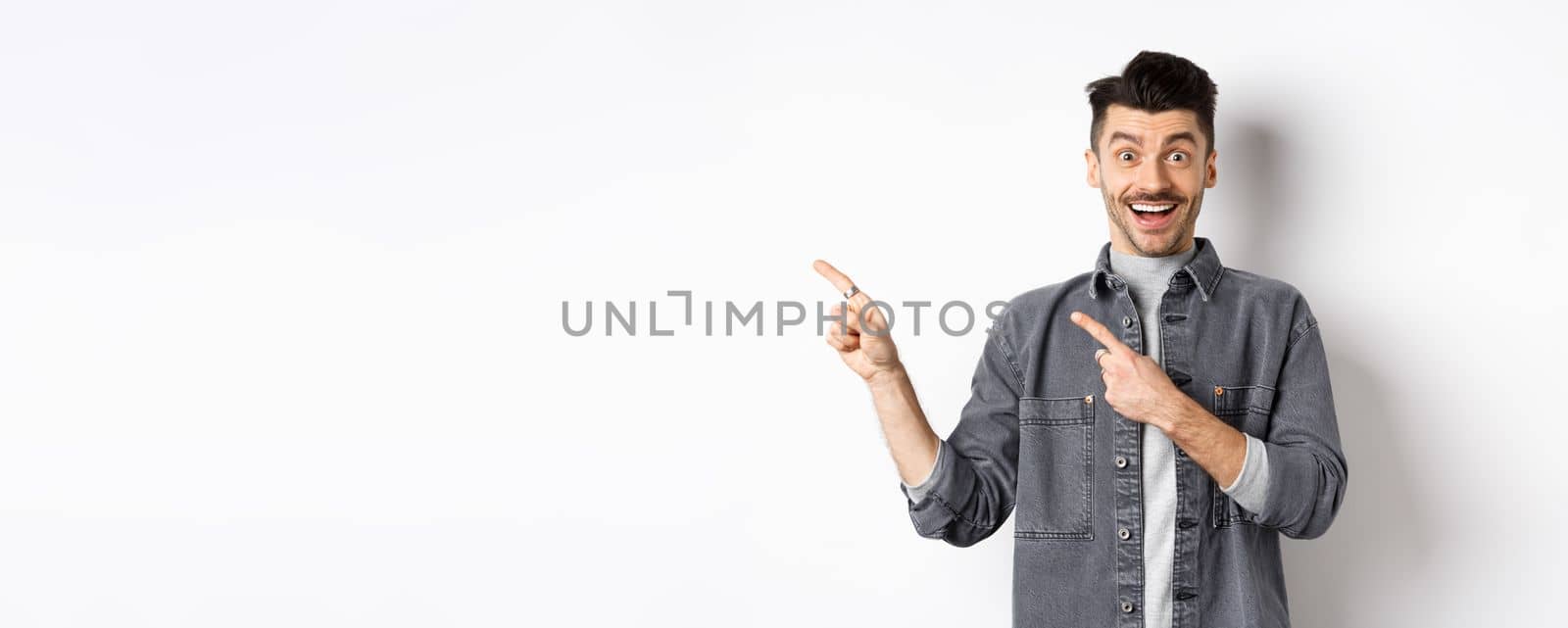 Excited guy with moustache pointing fingers left, showing advertisement and smiling happy, standing on white background.