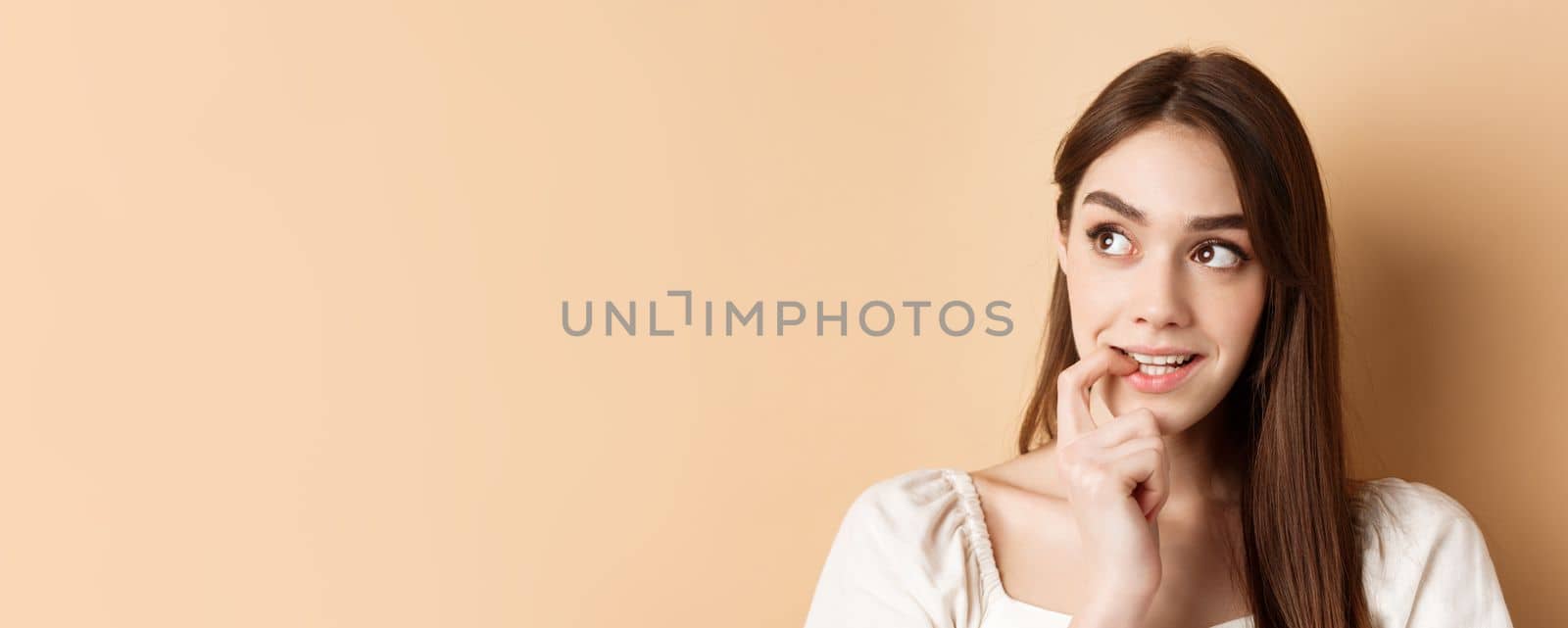 Close up of cute young woman thinking, biting fingernail and look tempted aside, staring at logo with desire, standing on beige background.