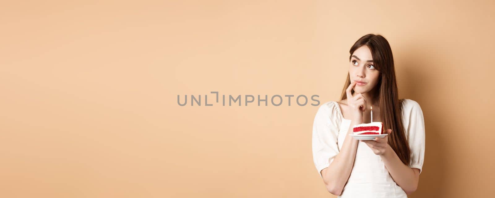 Dreamy birthday girl look aside and thinking of wish, blowing candle on cake, standing on beige background by Benzoix