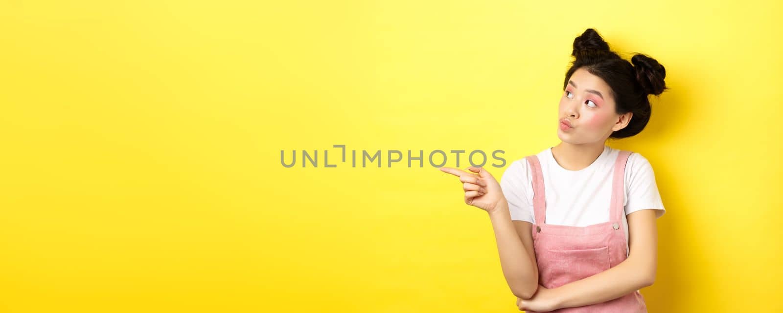Pensive asian woman with beauty makeup, pointing and looking left with curious face, interested in advertisement, standing on yellow background by Benzoix