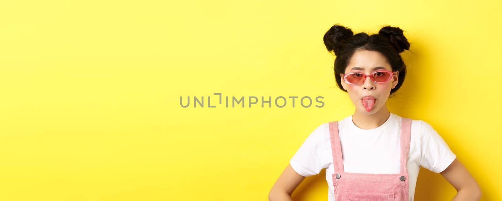 Summer and fashion concept. Silly asian teen girl showing tongue, wearing sunglasses, standing on yellow background by Benzoix