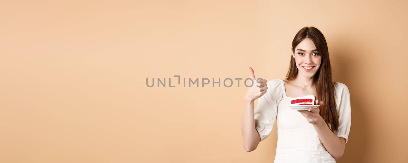 Beautiful birthday girl making wish on cake, showing thumb up and smiling happy, enjoying b-day party, standing on beige background.