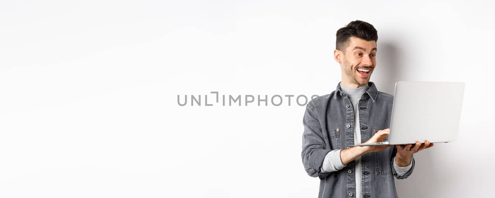 Man watching funny video on laptop, laughing at computer screen and smiling joyful, standing on white background by Benzoix
