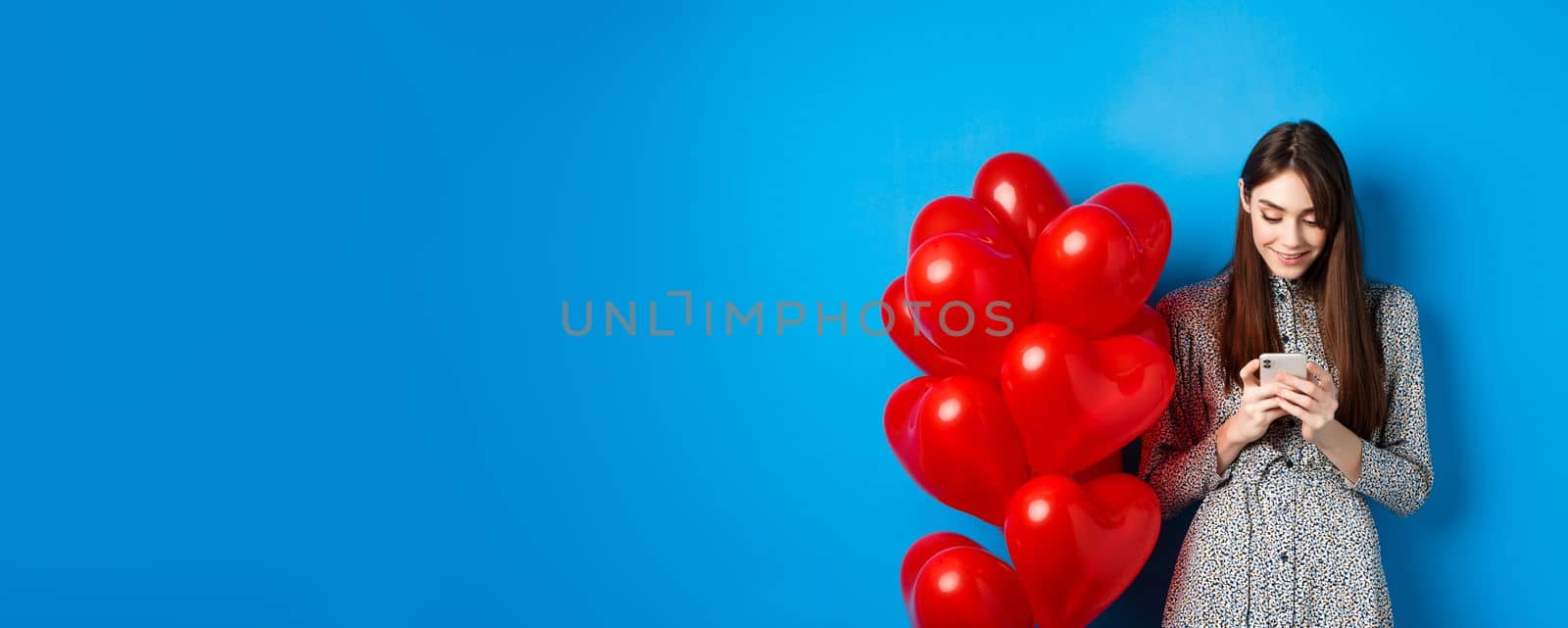 Valentines day. Smiling woman in dress standing near red hearts balloons and looking at smartphone, standing on blue background.