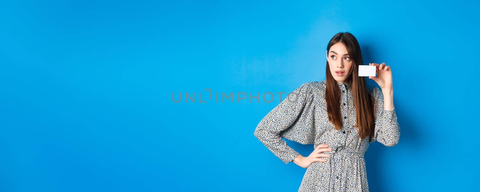 Shopping. Thoughtful female shopper looking left at logo, showing plastic credit card, standing on blue background by Benzoix