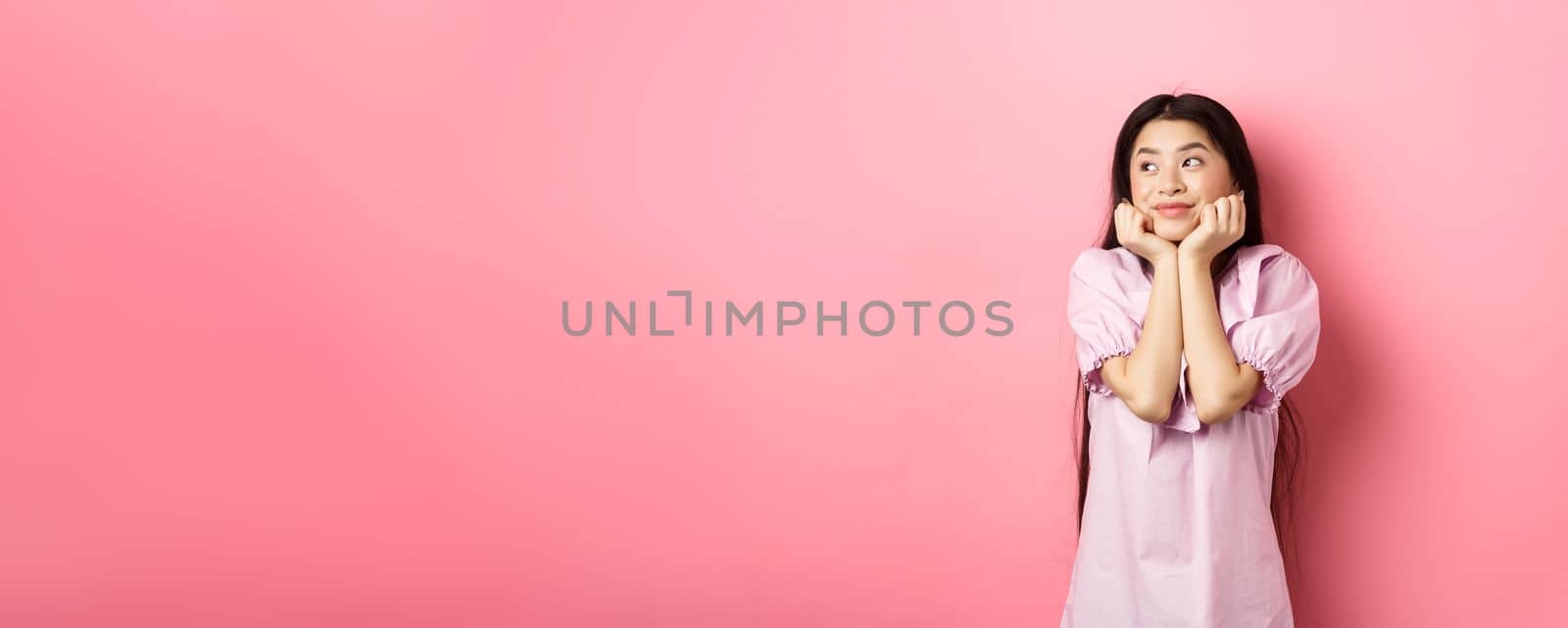 Dreamy romantic girl looking aside at logo and smiling, thinking of something beautiful, standing on pink background.