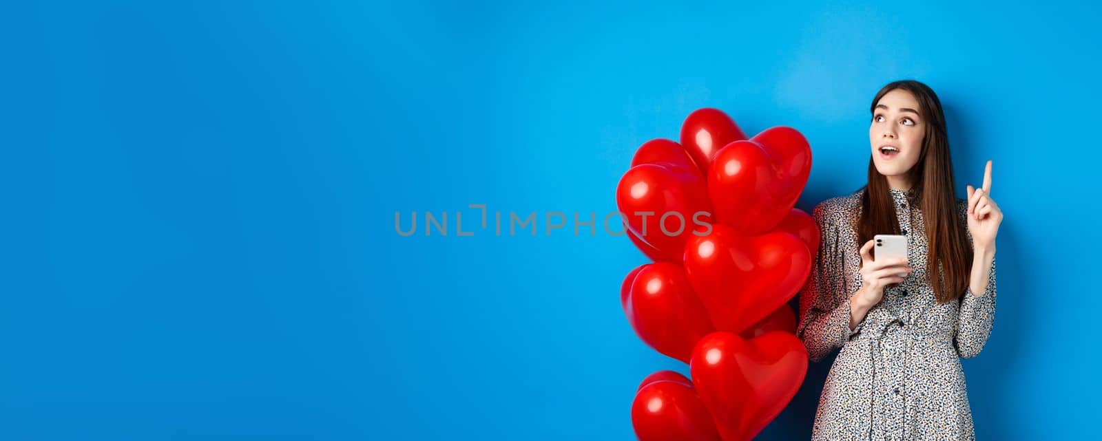 Valentines day. Image of romantic girl pitching an idea after using mobile phone, raising finger up and looking at empty space, standing near red heart balloons, blue background.