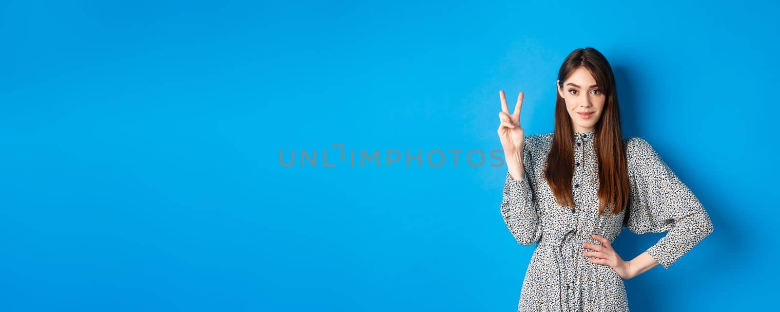 Young beautiful girl in dress with natural long hair, showing number two with fingers and smiling, standing against blue background by Benzoix