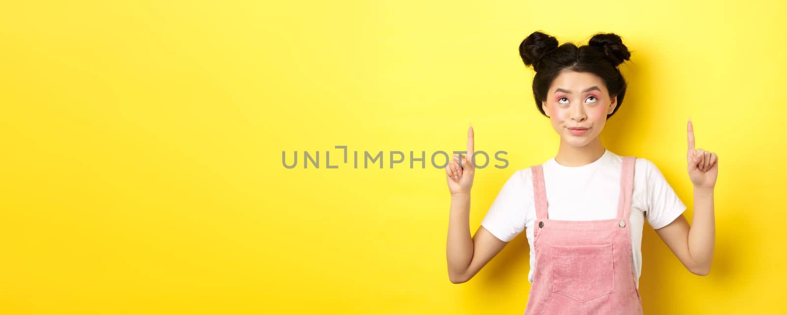Glamour asian girl with beauty makeup, pointing fingers up, showing top advertisement, standing on yellow background by Benzoix