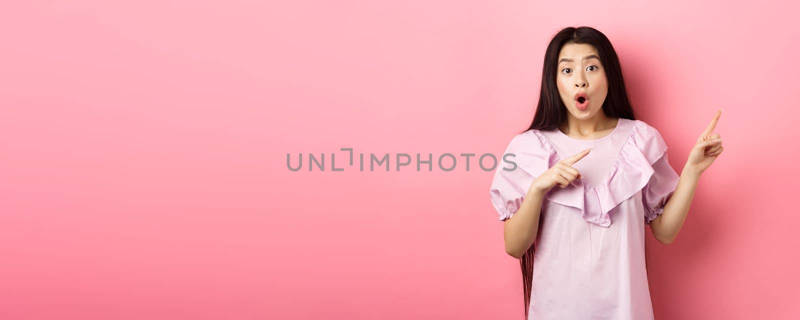 Excited asian woman look amazed, pointing right and gasping fascinated, checking out advertisement, standing on pink background.
